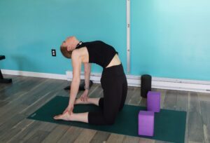 Dr. Chloe doing camel pose. She is on her knees with her feet straight behind her with her hips and spine extended forward in a backbend reaching for her heels behind her.